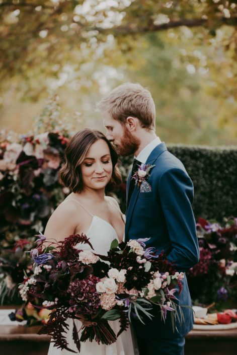 Bride holding bouquet desinged with burgundy hydrangea and amaranthus and dahlia, blush carnations and spray roses and groom with boutonniere