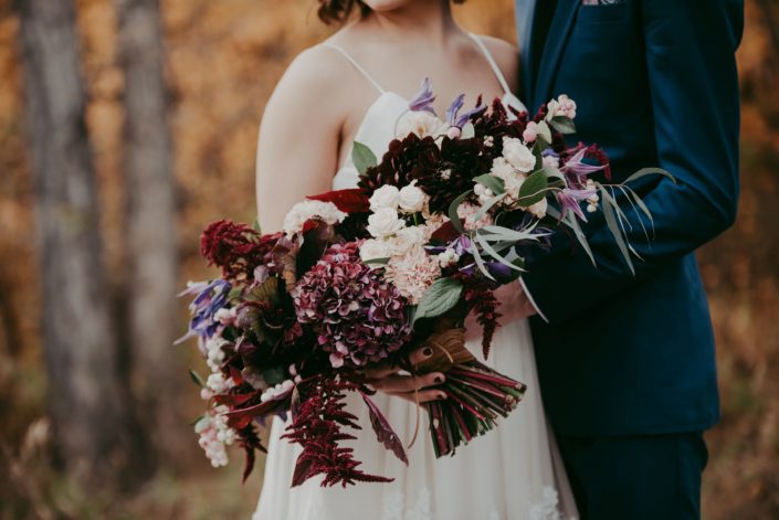 Close up of bridal bouquet designed with burgundy amaranthus and hydrangea blush carnations and spray roses and purple clematis with groom in navy blue suit