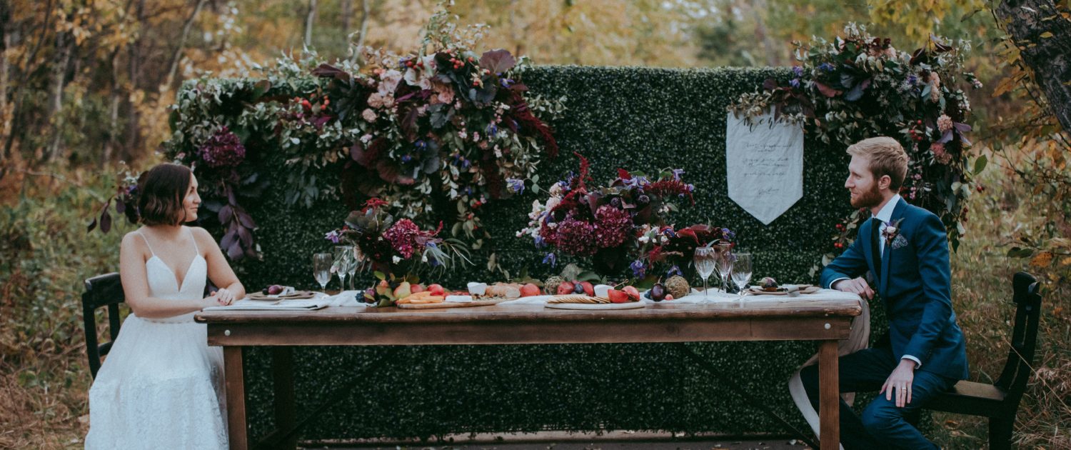 Styled shoot at McKenzie trails in Red Deer with bride and groom sitting at a tabe with a boxwood backdrop covered in autumn florals and charcuterie board