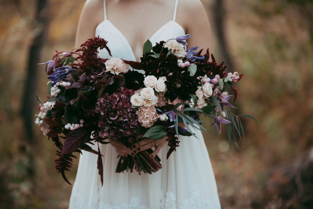 Bride in simple white dress holding a front facing bride bouquet designed with burgundy maranthus, hydrangea, blush spray roses, blush carnations and purple clematis for an autumn wedding
