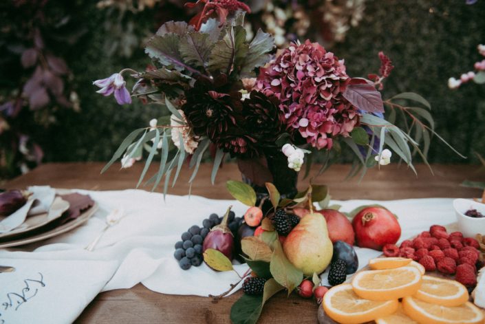 Fresh fruit on harvest table with charcuterie board and floral arrangement of burgundy dahlia, hydrangea and kale with purlpe clematis accents and snowberry