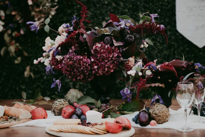 Boxwood wall backdrop with rustic harvest table with floral arrangement in a black compote with burgundy amaranthus and hydrangea, plum scabiosa, blush snowberries and purple carnations and fresh charcuterie boards