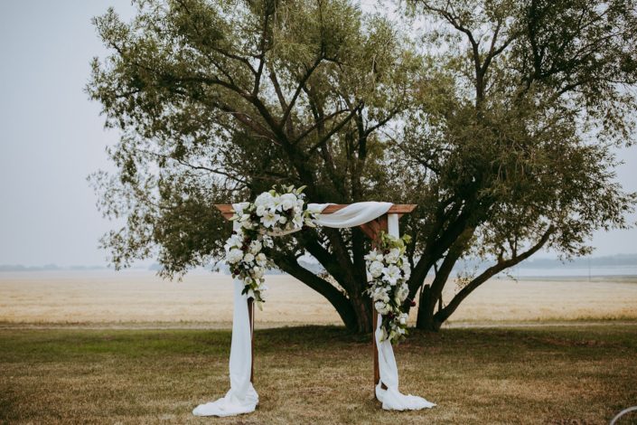 summer wedding archway with giant tree in wheat field with flowers in white including lilies and hydrangea and roses and burgundy dahlia