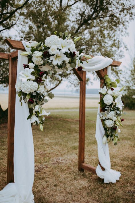 rustic archway with white voile and flower arrangements made with white hydragea and white lilies and roses and burgundy dahlia