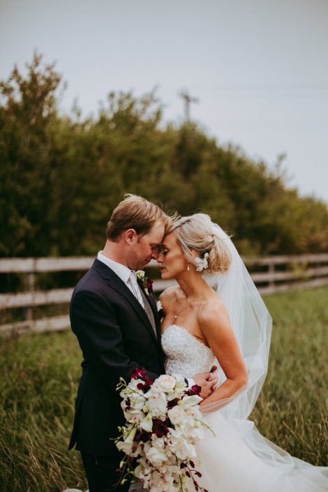 bride and groom in a summer wheat field in a alberta farm wedding holding a bridal bouquet of white and burgundy flowers including burgundy dendrobium and cymbidium orchids