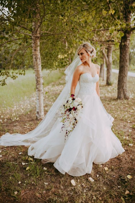 Bride in the trees in a summer wedding wearing a veil and holding a cascade bridal bouquet designed with dendrobium orchids and cymbidium orchids in white and burgundy and white calla lilies
