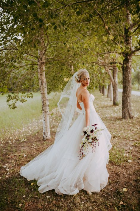 bride in the trees in a summer wedding with a veil holding a bouquet of white calla lilies and white dendrobium orchids and burgundy dendrobium orchids