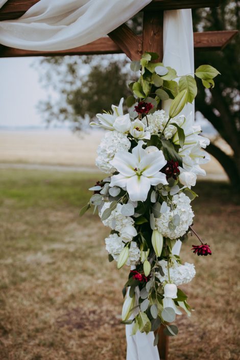 close up photo of archway arrangment with white hydrangea and white lilies and burgundy dahlia