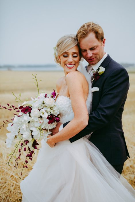bride and groom in alberta summer wheat field holding a modern cascade bridal bouquet designed with white dendrobium orchids, burgundy dendrobium orchids and white roses
