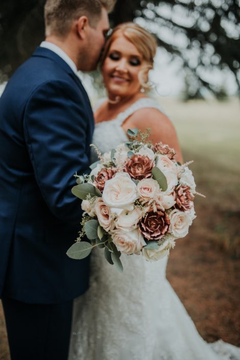Close up of bridal bouquet made with rose gold succulents white ohara garden roses and blush quicksand roses and eucalyptus held by bride and groom in navy suit