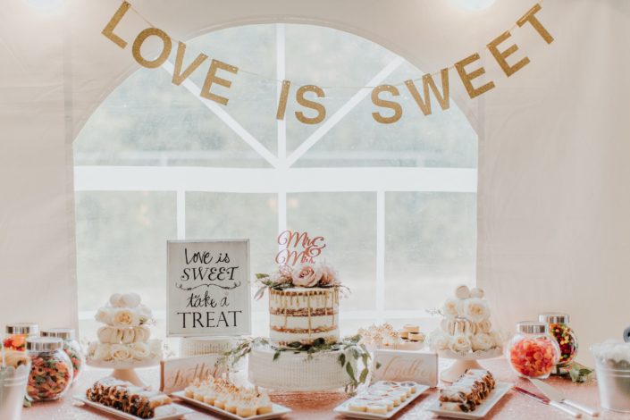 Dessert table set with naked cake and rose gold accents with candy bar and small desserts and love is sweet banner
