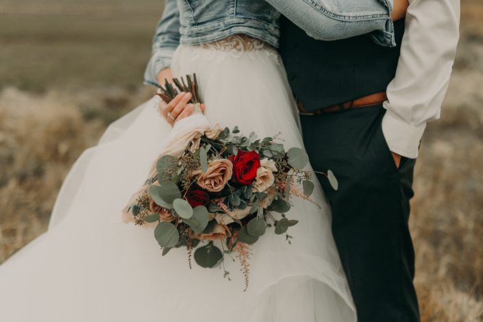 country bride and groom in prairie field with jean jacket on holding a bouquet with red roses and white o'hara garden roses and quicksand roses and eryngium and pink astilbe and eucalyptus
