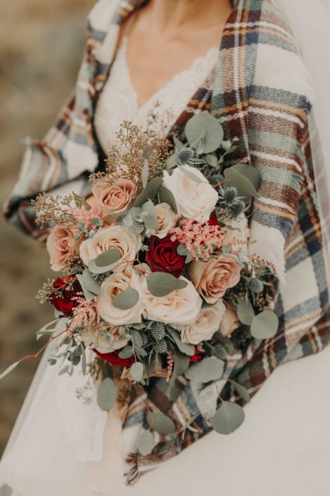 bride in plaid shawl with bouquet with red roses and quicksand roses and white o'hara garden roses and astilbe and eryngium and eucalyptus
