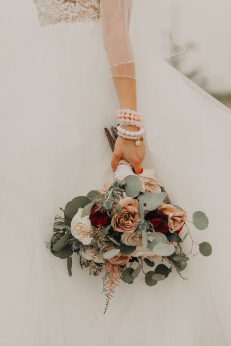 bride with lace dress and stacked bracelets holding a bouquet with quicksand roses and red roses and white o'hara garden roses and pink astilbe and eryngium and seeded eucalyptus and silver dollar eucalyptus