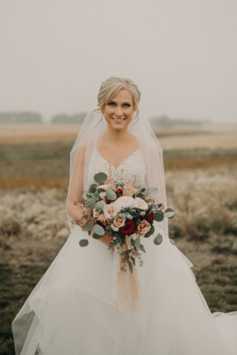 bridal portrait of bride with veil and lace dress with blush bouquet with quicksand roses and white o'hara garden roses and pink astilbe and red roses and blue eryngium and silver dollar eucalyptus in a prairie field