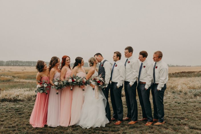 bridal party with kissing bride and groom with bridesmaids in various blush colored dresses and groomsmen in grey pants with blue ties and red spray rose boutonnieres and bouquets with red roses and quicksand roses and white o'hara garden roses and pink astilbe and mixed eucalyptus