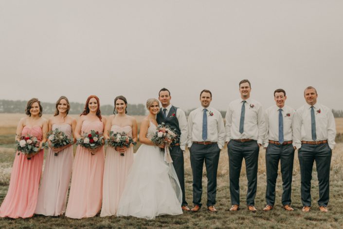 bridal party with bridesmaids in blush dresses of various shades and holding bouquets with white and red and blush roses and eucalyptus groomsmen in grey pants with blue ties and boutonnieres with red spray roses