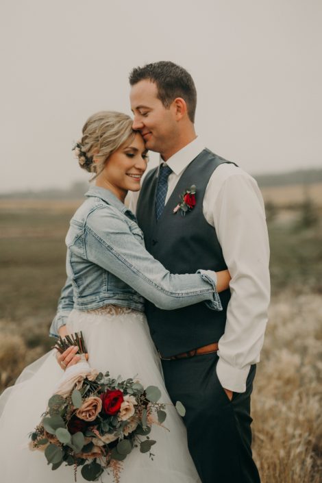 bride in jean jacket with groom in grey suit with red spray rose boutonniere and bouquet with blush and red roses and pink astilbe and eucalyptus in prairie field