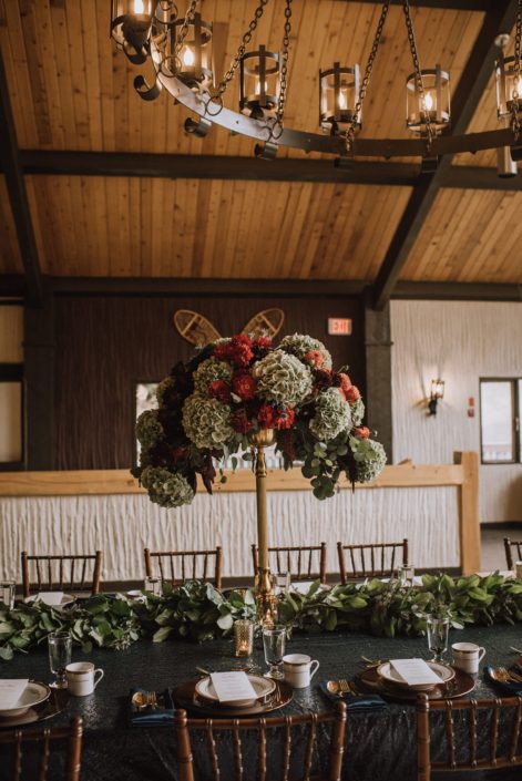 A tall centerpiece designed atop a gold candleabra and eucalytus table runner placed along a live edge table at Canyon Ski Resort long table dinner