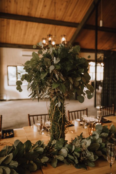 Tall centerpiece with moss covered trunk and mixed eucalyptus topper accented with curly willow and a greenery garland runner on a live edge table with wood chairs in canyon ski resort in Red Deer Alberta