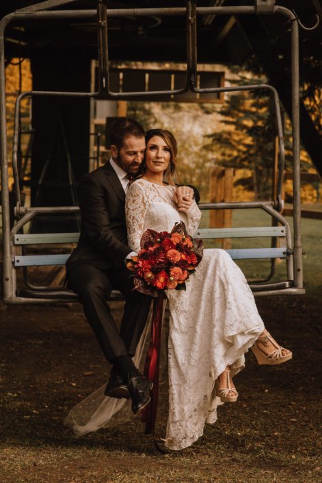 bride and groom on ski lift with lace dress in fall and bouquet with orange dahlia and red roses and red dahlia and burgundy anthurium and magnolia leaves and trailing ribbons