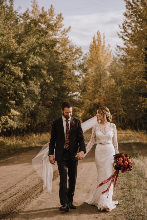 Bride in long sleeved dress with long veil with groom in floral tie walking down a dirt road in the woods with a bouquet with trailing ribbons and orange dahlias and black bacarra roses and burgundy anthuriums and magnolia leaves