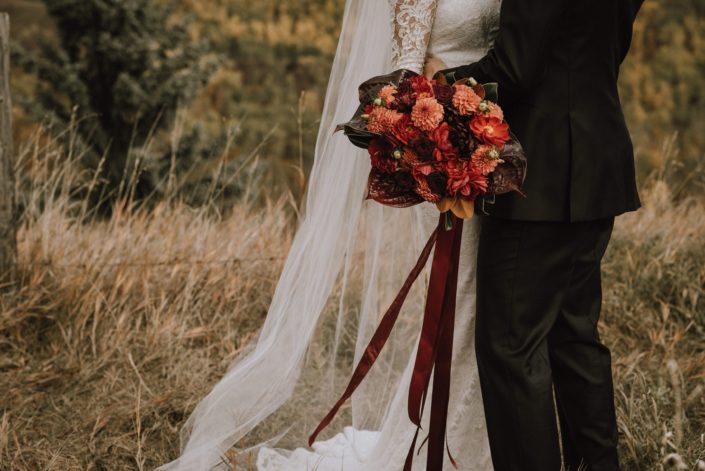 bride and groom in rustic field with lace dress and bouquet with orange dahlia and black bacarra roses and anthuriums and magnolia leaves and trailing ribbons