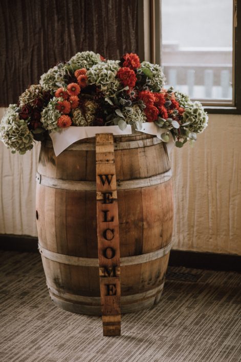 Rustic wedding welcome arrangement with welcome sign and flowers including green hydrangea and orange dahlia and red dahlia and red roses and blue eryngium