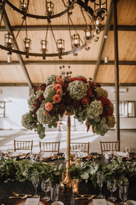 tall centerpiece on top of a gold candelabra with green hydrangea and red dahlia and red roses and orange dahlia accented by a mixed greenery garland on the long table at canyon ski resort in Red Deer Alberta
