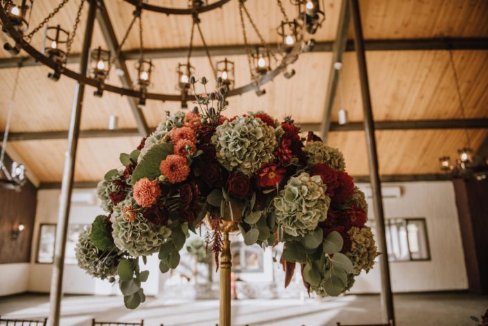 Breathtaking tall centerpiece atop a gold candelabra with green hydrangea and orange dahlias and red dahlias and eucalyptus and eryngium and red roses at Canyon Ski Resort in Red Deer Alberta
