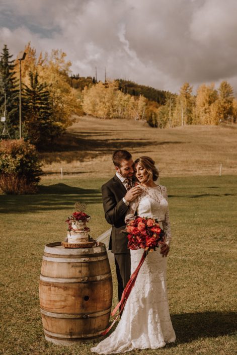 Bride and Groom at Canyon Skir Resort in the autumn holding a Dahlia bouquet in burgundy and burnt orange standing beside a rustic whiskey barrel with a naked wedding cake on it