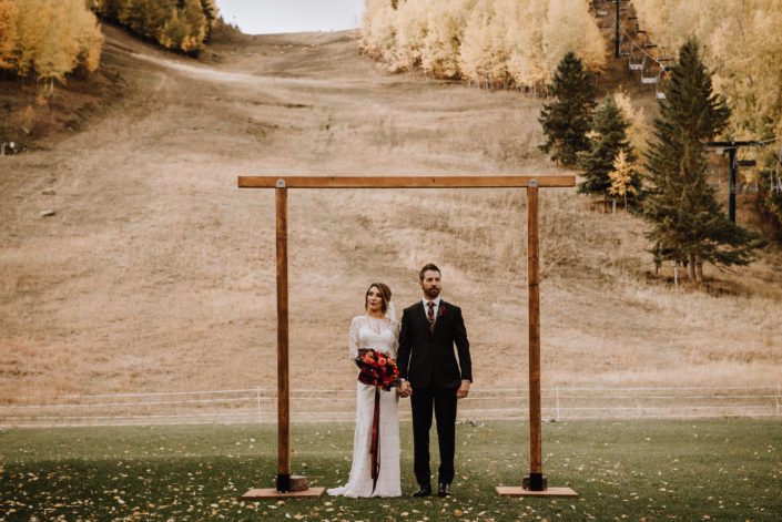 Bride and Grooms stand under a wooden archway at Canyon Ski Resort in the autumn Bride is holding a bouquet of dahlias in orange and burgundy