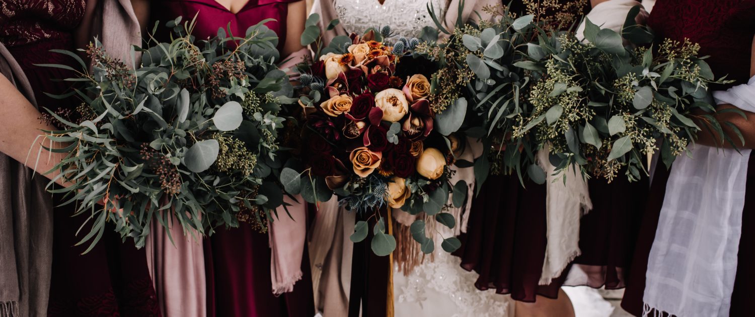 Bride and bridesmaids in deep burgundy holding bouquets designed with mixed eucalyptus greenery, burnt orange chrysanthemum, peach garden roses, burgundy roses and burgundy dahlia