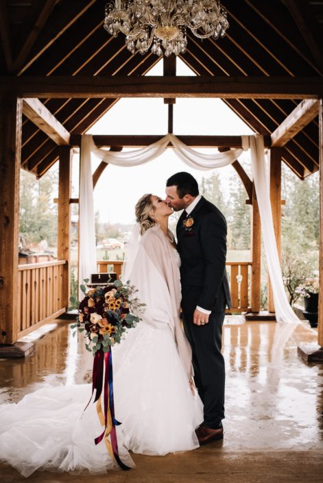 bride and groom kissing at the pavillion at meridian beach in the autum holding a bridal bouquet designed with mustard yellow chrysanthemum and garden roses, burgundy roses and eucalyptus