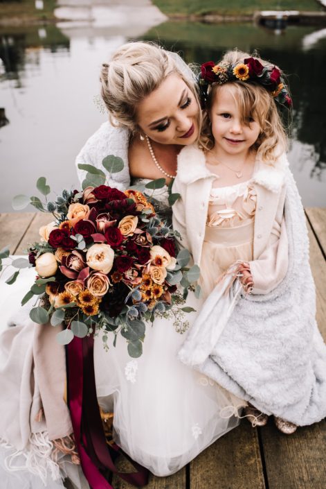 bride and flowergirl in white for a rustic autumn wedding on a lake front holding a bridal bouquet of mustard yellow garden roses, burgundy spray roses and peach roses with santini chrysanthemums and silk trailing ribbons and a flowercorwn