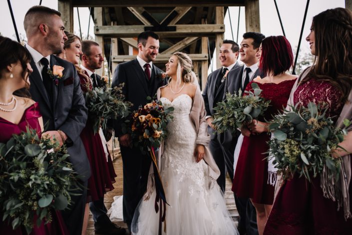 Wedding party with bride, groom bresmaids and groomsmen standing on a rustic bridge holding eucalyptus grenery bouquets and bridal bouquet in autumn colors