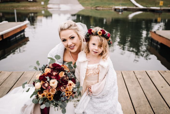 Bride with a bridal bouquet designed with burgundy roses, peach garned roses, burnt orange chrysanthemum and eucalyptus with a flowergirl wearing a flowercrown of red spray roses and chrysanthemum