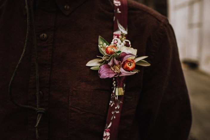 alternative groom in floral suspenders with boutonniere with red ranunculus buds and purple clematis