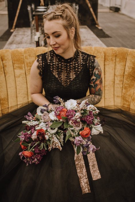 alternative bride in black wedding dress with tattoos on a yellow sofa with a bouquet of red ranunculus and amnesia roses and white anemone and white pieris and plum scabiosa and clematis