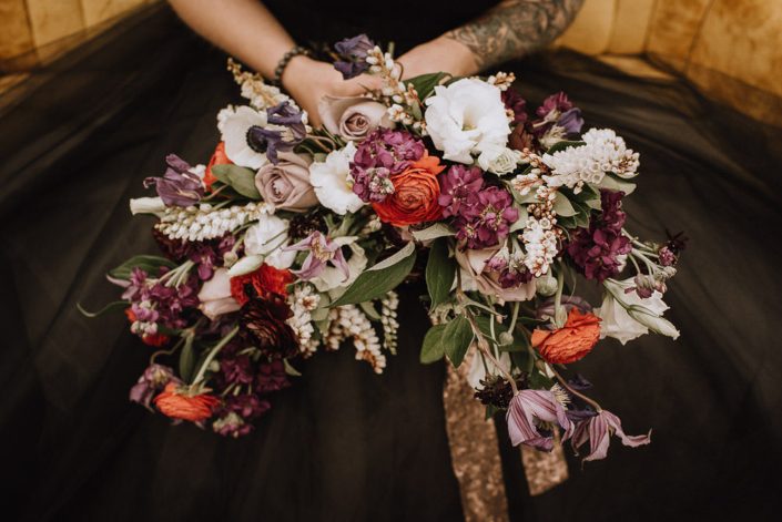 alternative bridal bouquet with black wedding dress and bouquet with red ranunculus and purple stock and white lisianthus and purple clematis and plum scabiosa and amnesia roses and a copper sequin wrap