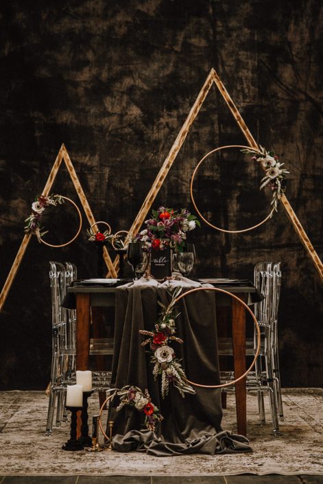 alternative wedding backdrop with acrylic chairs and black backdrop and floral hoops with white anemone and red ranunculus and white pieris and clematis and triangle archways and a wood table