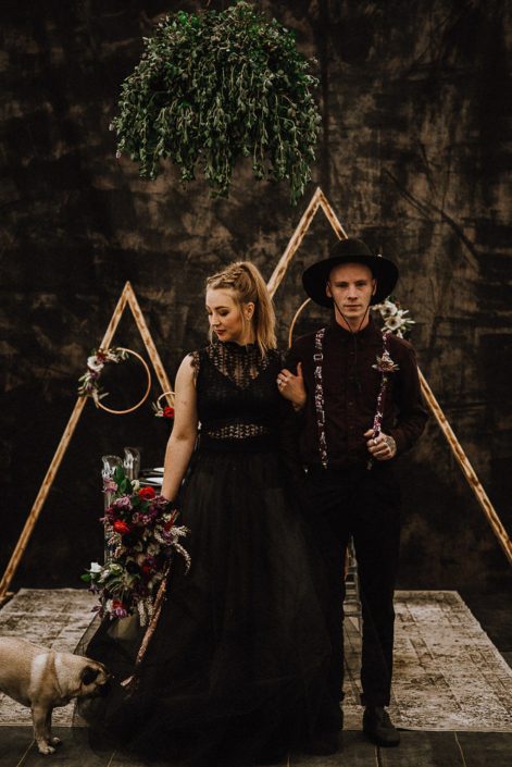 alternative wedding photo with black bridal dress and groom in black with suspenders and wide brim hat with triange arch backdrop and bouquets with red and burgundy and floral hoops and area rugs and pugs