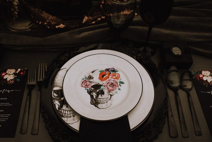 Alternative wedding table setting with skulls and floral plates and black flatware and black chargers and grey velvet runner