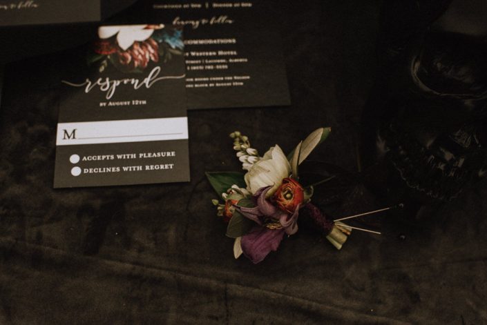 boutonniere close up with red ranunculus and purple clematis and white pieris and white anemone on velvet cloth with black invitation in background