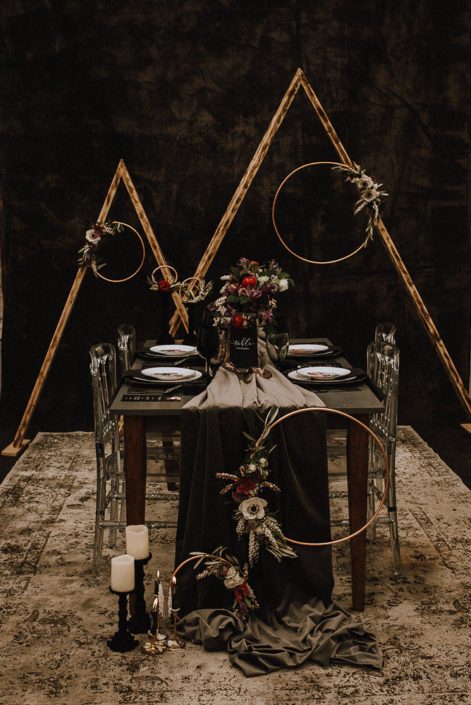 Alternative wedding decor dark and moody black backdrop with triangle arches and copper floral hoops ad grey velvet table runner on a wood table with acrylic chairs and red ranunculus and white anemone and black chargers