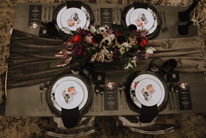 alternative wedding table setting with flower skull plates and black chargers and grey velvet table runner and centerpiece with red ranunculus and white anemone and amnesia roses and purple clematis and white lisianthus and white pieris