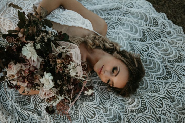 bride laying on lace dress with boho style braid in her hair and bridal bouquet of blush roses and burgundy leaves