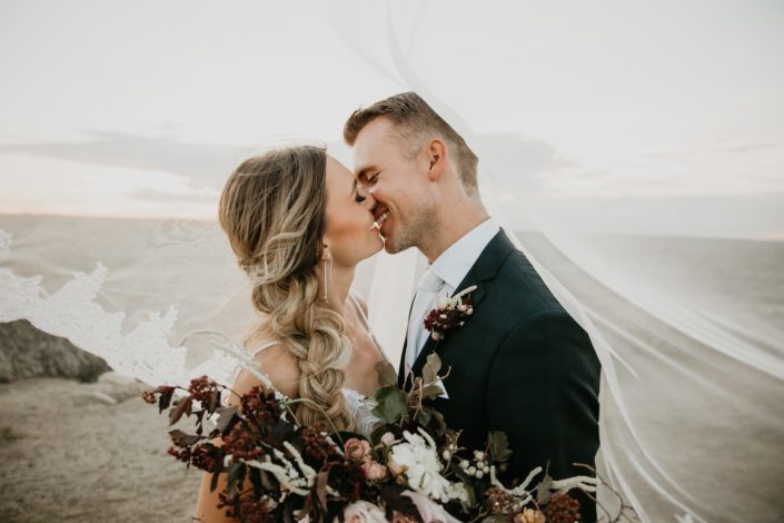 bride and groom kissing under her veil with bride holding an ivory and burgundy floral bouquet
