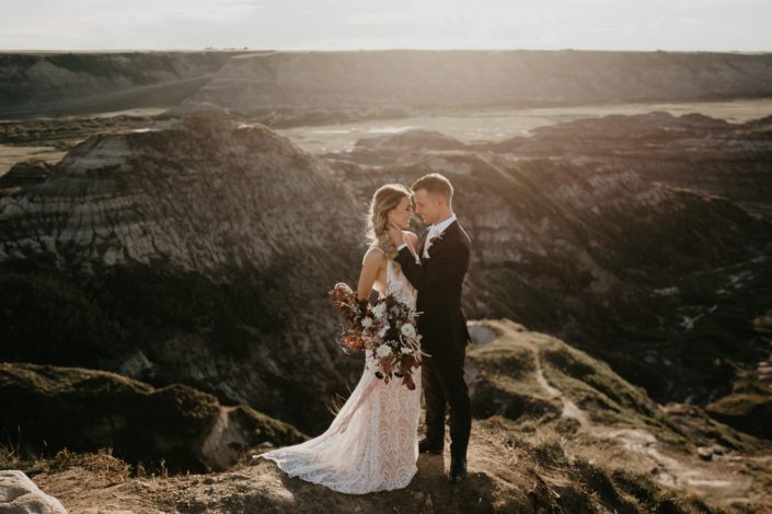 bride in a lace dress holding an organic bridal bouquet and groom overlooking the badlands in drumheller at sunset