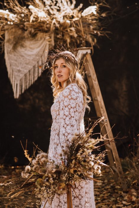 fall boho bride with dried floral bouquet and boho floral fall crown in front of a pampas grass and dried floral archway piece with crochet in the woods